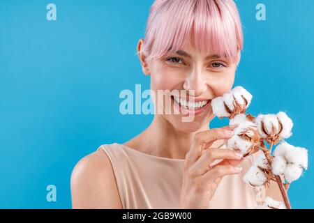 Porträt einer hübschen jungen Frau mit rosa Haaren, die an der Kamera lächelt und einen Zweig mit flauschigen Baumwollblumen hält und isoliert über dem blauen Studio posiert Stockfoto