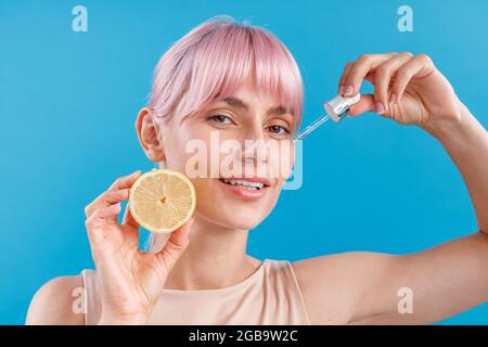 Porträt einer attraktiven Frau mit perfekter Haut und rosa Haaren, die eine Hälfte der frischen Zitrone und eine Pipette in der Nähe ihres Gesichts auf blauem Hintergrund isoliert hält Stockfoto