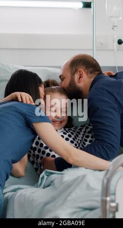 Glückliche Eltern besuchen kranke Tochter umarmen Kind während der Untersuchung auf der Krankenstation. Hospitalisiertes Kind mit Sauerstoff-Nasenschlauch, der nach einer Genesungsoperation im Bett ruht Stockfoto