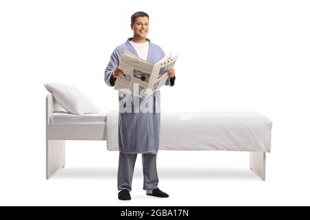 Junger Mann mit einer Zeitung, die einen Bademantel trägt und neben einem Bett auf weißem Hintergrund steht Stockfoto