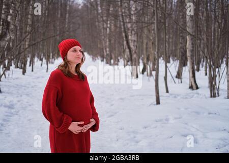 Eine glückliche Schwangere in einem Strickpullover und einem Hut spaziert im Park, einem Winterwald mit Bäumen im Schnee Stockfoto