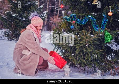 Eine glückliche Frau in Winterkleidung legt am Silvesterabend in einem verschneiten Wald Geschenke unter den Weihnachtsbaum Stockfoto