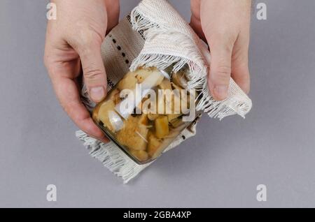 Ein Glas mit hausgemachten eingelegten Pilzen vor grauem Hintergrund. Ein Mann hält den Pappelpilz aus der Dose oder Tricholoma populiinum. Stockfoto