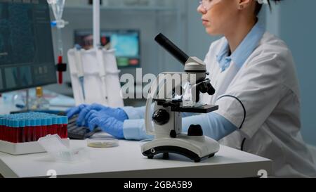 Nahaufnahme des Mikroskop-Werkzeugs, während Wissenschaftler Petrischale analysieren und modernen Computer im Hintergrund verwenden. Frau am Labortisch mit Blutprobe und Tablett mit Staubsaugern zur Untersuchung Stockfoto