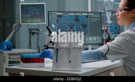 Wissenschaftlerin sitzt im Labor mit chemischer Technologie auf dem Schreibtisch. Biochemie Arzt mit Pipette, Blutvacutainser, Mikroskop, Eingabe am Computer für mikrobiologische Experiment Stockfoto