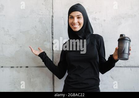 Junge muslimische Frau in Hijab lächelt, während sie Wasserflasche und Kopieplatz in Innenräumen hält Stockfoto