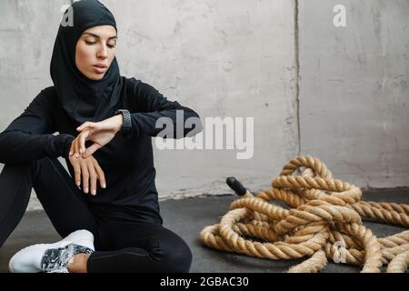 Junge muslimische Frau im Hijab, die Smartwatch anschaut, während sie drinnen auf dem Boden sitzt Stockfoto