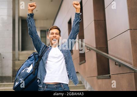 Mittellange Aufnahme eines glücklichen, hübschen jungen Mannes mit Lieferservice und großem Thermorucksack, der die Hände hebt und den Sieg feiert. Stockfoto