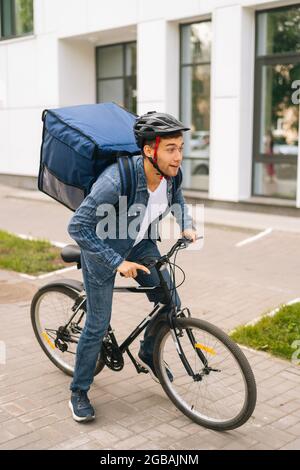 In voller Länge vertikale Porträt von fröhlich hübsch junge Lieferung Mann in Schutzhelm Start Fahrrad Fahrt in der Stadt Straße. Stockfoto