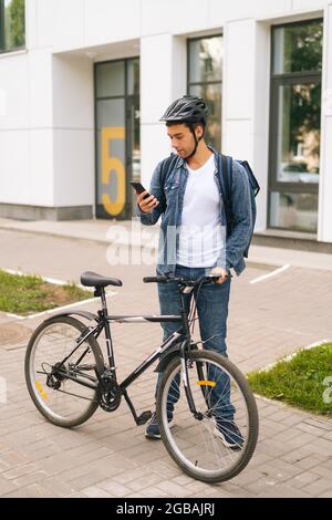 Porträt eines gutaussehenden männlichen Kuriers mit Thermorucksack, der mit dem Fahrrad auf der Stadtstraße steht und die Navigations-App am Telefon nutzt. Stockfoto