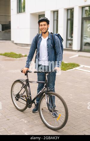 Vertikales Porträt von fröhlich hübsch junge Lieferung Mann in Schutzhelm posiert in der Nähe Fahrrad in der Stadt Straße. Stockfoto
