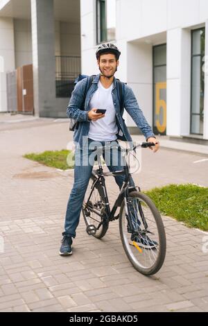 In voller Länge vertikale Porträt von fröhlichen männlichen Kurier mit Rucksack stehen mit Fahrrad in der Stadt Straße und mit Navigation App am Telefon. Stockfoto