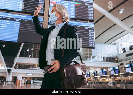 Geschäftsmann mit Gesichtsmaske am Flughafenterminal winkt jemanden an. Geschäftsreisende, die nach der Sperre von Covid-19 reisen. Stockfoto