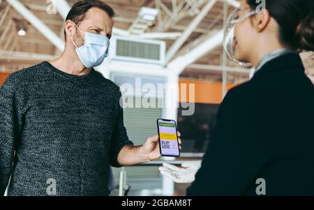 Männlicher Reisender in Gesichtsmaske, der dem Bodenwächter seinen Impfpass auf dem Smartphone zeigt. Flugbegleiter prüft Impfpass von Mann A Stockfoto