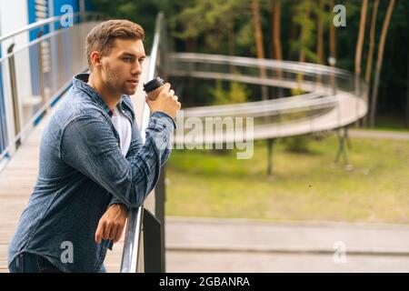 Seitenansicht eines selbstbewussten, nachdenklichen jungen Mannes, der auf der Terrasse des Balkons stand und Kaffee am Geländer des Bürogebäudes trank. Stockfoto