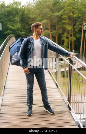 In voller Länge vertikale Porträt von schönen jungen Mann der Lebensmittelzustellung Service mit großen thermischen Rucksack auf der Terrasse des Balkons stehen. Stockfoto