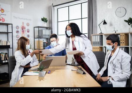 Zwei Männer und zwei Frauen in Laborkittel mit Stethoskopen sitzen am Tisch und verwenden moderne Gadgets, afro Dame, die Papiere für Kollegen. Multirassische Ärzte analysieren die Diagnose eines schweren Patienten. Stockfoto