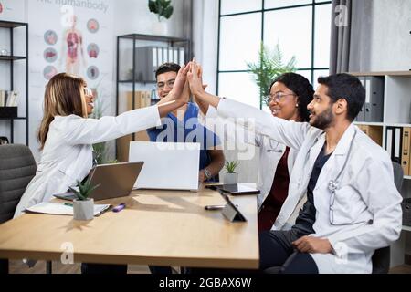 Team von vier internationalen Ärzten, die fünf hohe für die erfolgreiche Zusammenarbeit bei gemeinsamer Arbeit und medizinischen ptoject Ausdruck. Mitarbeiter im Gesundheitswesen sitzen zusammen an Tablets und verwenden moderne Geräte. Stockfoto