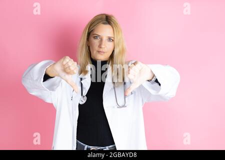 Junge blonde Ärztin, die Stethoskop trägt und auf einem isolierten rosa Hintergrund steht und dabei das negative Zeichen mit dem Daumen nach unten zeigt, dass sie nicht gefällt Stockfoto