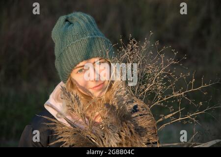 Porträt eines jungen schönen Mädchens in einem warmen Strickmütze und Jacke. Hält im Herbst ein Bouquet aus trockenem flauschigen Schilf, beleuchtet von Sonnenlicht, lächelnd Stockfoto