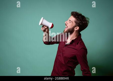 Bärtiger junger Mann, der in einem Megaphon laut redet. Konzept von Menschen, die ihre Stimme über die Menge hinaus erheben. Stockfoto