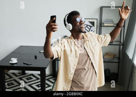 Glücklicher afrikanischer Mann mit kabellosen Kopfhörern, der sein Mobiltelefon benutzt, um Musik zu hören und im Zimmer zu tanzen Stockfoto