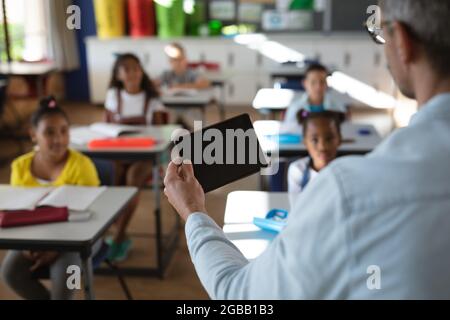 Rückansicht des kaukasischen männlichen Lehrers, der während des Unterrichts in der Schule ein digitales Tablet verwendet Stockfoto