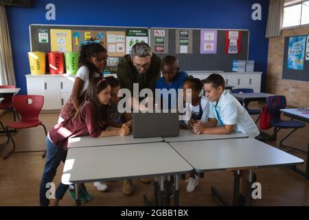 Kaukasischer Lehrer und eine Gruppe verschiedener Schüler, die in der Schule gemeinsam einen Laptop benutzen Stockfoto