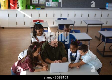 Kaukasischer Lehrer und eine Gruppe verschiedener Schüler, die in der Schule gemeinsam einen Laptop benutzen Stockfoto