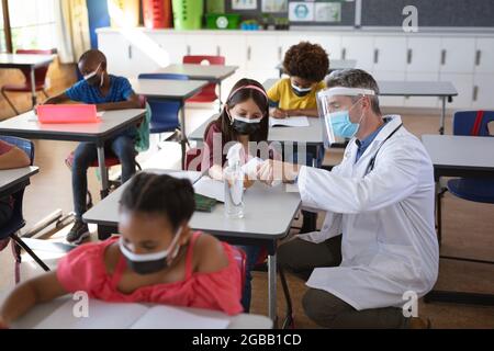 Kaukasischer Arzt mit Gesichtsschutz, der die Temperatur des kaukasischen Mädchens in der Schule misst Stockfoto