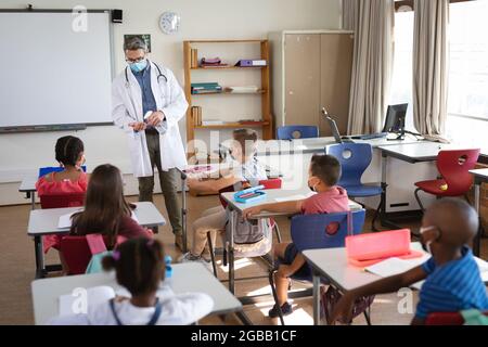 Kaukasischer Arzt mit Gesichtsmaske zeigt, wie man Händedesinfektionsmittel für Schüler in der Schule verwendet Stockfoto
