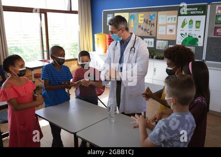 Kaukasischer Arzt mit Gesichtsmaske zeigt, wie man Händedesinfektionsmittel für Schüler in der Schule verwendet Stockfoto