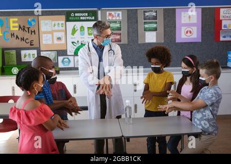 Kaukasischer Arzt mit Gesichtsmaske zeigt, wie man Händedesinfektionsmittel für Schüler in der Schule verwendet Stockfoto