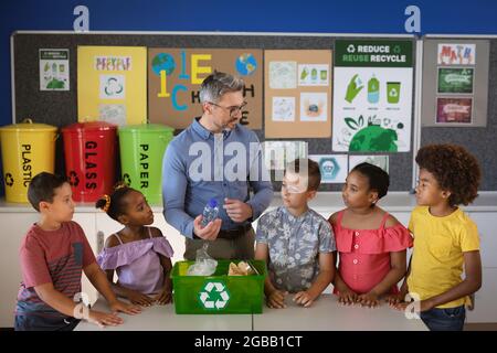 Kaukasischer männlicher Lehrer, der eine Gruppe verschiedener Schüler unterrichtet, um Plastikgegenstände in der Schule zu recyceln Stockfoto