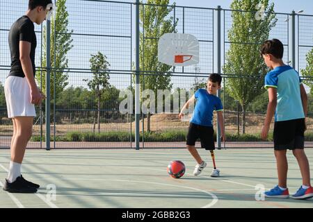 Couch, die mit zwei Schülern Fußball spielt. Einer von ihnen hat eine Beinprothese und tritt den Ball. Stockfoto