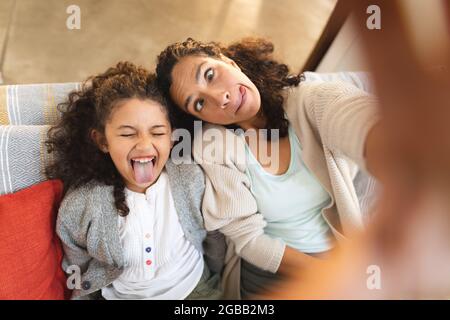 Mutter und Tochter mit gemischter Rasse sitzen auf dem Sofa und machen lustige Gesichter, machen Selfie Stockfoto