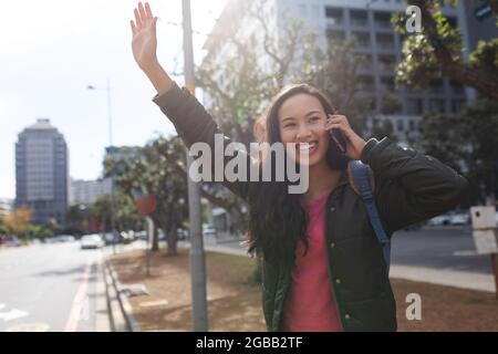 Asiatische Frau, die auf der Straße steht und ein Taxi anhagelt und auf dem Smartphone spricht Stockfoto