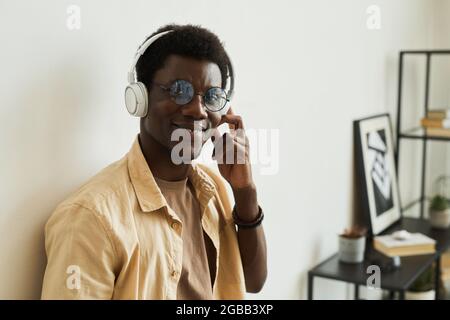 Porträt eines afrikanischen jungen Mannes in einer Brille, der Musik in kabellosen Kopfhörern hört und die Kamera anlächelt, während er im Zimmer steht Stockfoto
