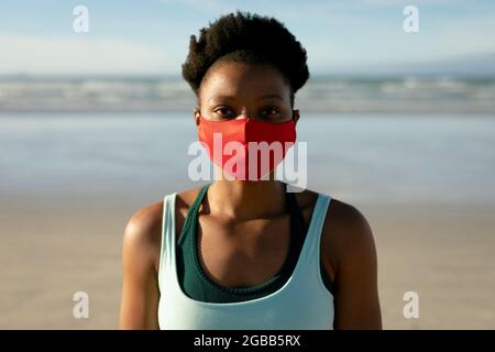 Portrait einer afroamerikanischen Frau, die eine Gesichtsmaske trägt und Yoga praktiziert, am Strand steht Stockfoto