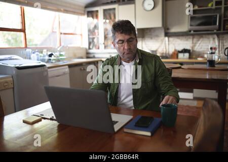 Kaukasischer Mann in der Küche, der mit einem Laptop am Tisch sitzt und Notizen schreibt Stockfoto