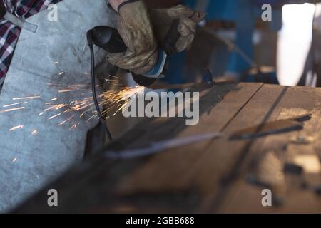 Hände des kaukasischen männlichen Messerherstellers, der Schürze trägt und Winkelschleifer in der Werkstatt verwendet Stockfoto