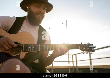 Junger bärtiger Mann mit Hut, der am sonnigen Tag Gitarre auf dem Dach spielt Stockfoto