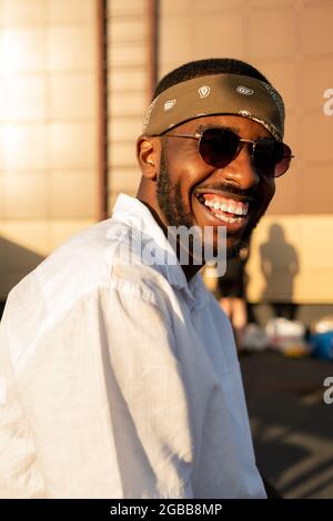 Fröhlicher afrikanischer Kerl in weißem Hemd, Sonnenbrille und Stirnband genießen Party im Freien Stockfoto
