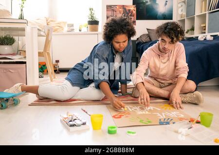 Interkulturelles Teenager-Paar, das Plakat auf ein Stück Pappe zeichnet, während es auf dem Boden sitzt Stockfoto