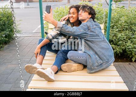 Unbeschwerte Teenager-Dates machen Selfie auf Schaukeln, während Kerl umarmt seine glückliche Freundin Stockfoto