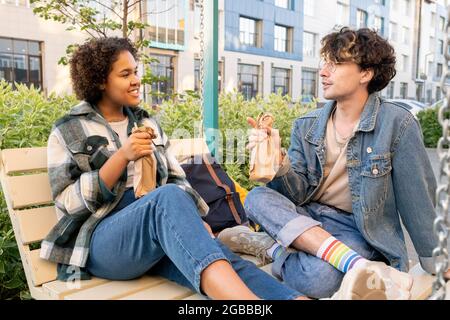 Junges interkulturelles Paar in Casualwear, das beim Entspannen auf Schaukeln im Freien Getränke trinkt Stockfoto