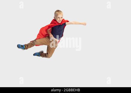 Porträt eines kleinen blonden Jungen, Schüler fliegen wie ein Superheld in rotem Umhang isoliert auf weißem Studiohintergrund. Copyspace für Werbung. Kindheit, Bildung Stockfoto