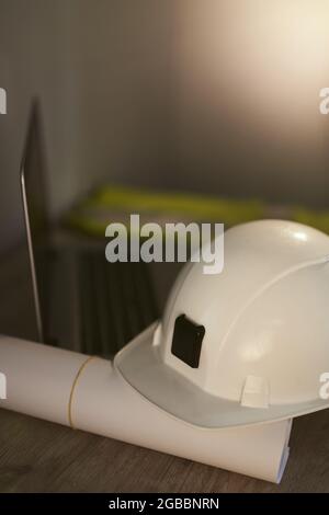 Weißer Schutzhelm, gerollte Entwürfe oder Pläne und Laptop-Computer auf dem Arbeitstisch des Vorgesetzten. Bau-, Architektur-, Design- oder Bauindustriekonzept. Hochwertige vertikale Fotos Stockfoto