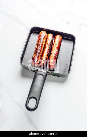 Schweinswürste, die in einer kleinen Antihaft-Bratpfanne auf einem Marmorhintergrund gekocht werden – FOODPIX WEISS Stockfoto