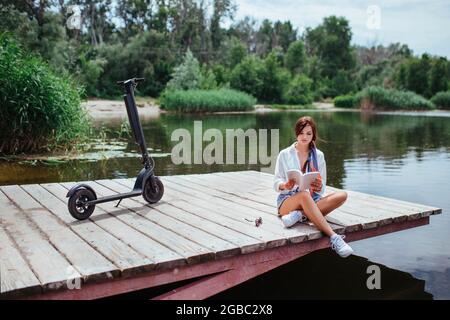 Ein schönes Mädchen mit einem Elektroroller liest ein Buch auf einer Holzbrücke am Fluss. Öko-Transport und Gesundheitskonzept. Stockfoto